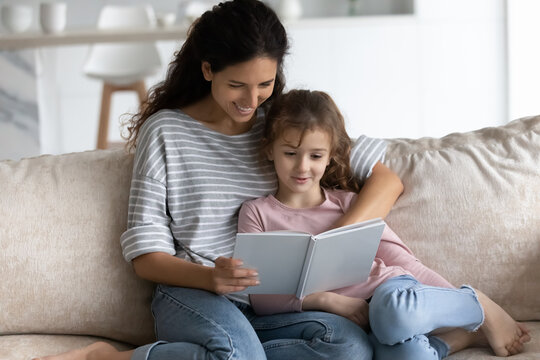 Smiling loving young Latino mother and small teen daughter sit relax on couch at home read book together. Happy caring Hispanic mom and biracial teenage girl child rest enjoy story in textbook.