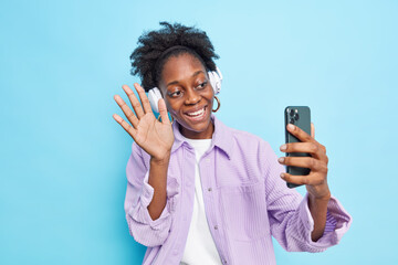 Positive dark skinned curly haired woman waves palm in hello gesture makes video call greets friend from abroad holds smartphone smiles broadly wears stylish purple shirt isolated over blue background