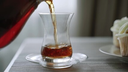 Black tea is poured from teapot into tall glass transparent cup, close-up. Morning homemade tea. Fresh appetizing cakes are nearby.
