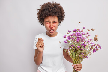 Allergy to pollen. Frustrated emotional Afro American woman frowns face keeps eyes closed reacts on trigger suffers fro allergy on pollen holds wildflowers. Spring exacerbation of allergies.