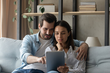 Young couple sit on sofa spend carefree weekend time at home using tablet device. Family having fun using new mobile application, buying on-line, choose services goods on internet, make order concept