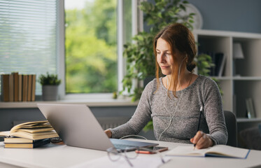 Beautiful woman in earphones sitting at living room and using modern laptop for for online education. Focused female taking notes while studying at home.