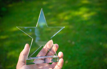 The hand holds a transparent star made of glass on a summer green background, the winner's cup, a...