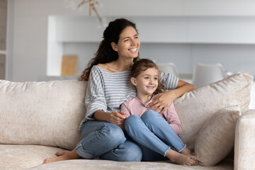 Obraz premium Happy Latino mother and biracial teen daughter sit relax on couch in living room cuddle and hug. Smiling young Hispanic mom and ethnic girl child rest on sofa at home look in distance dream embrace.