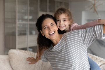 Obraz na płótnie Canvas Portrait of happy small ethnic girl child piggyback play with smiling Hispanic young mother relax together at home. Overjoyed Latino mom and teen daughter feel playful rest involved in funny game.