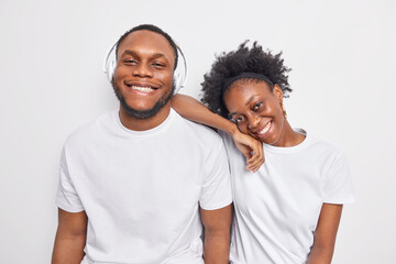 Positive friendly dark skinned woman and man happy to spend free time together smile pleasantly dressed casually listen music in headphones isolated over white background. Friendship concept