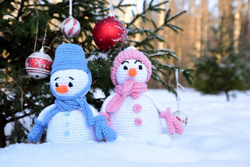 Knitted snowmen in colorful clothes stand in the snow under a Christmas tree decorated with toys