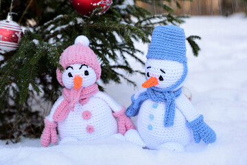 Two knitted Christmas snowmen, a boy and a girl, stand under a decorated Christmas tree outside