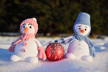 Two knitted snowmen stand under the bright winter sun in the snow. Between them lies a Christmas tree ball