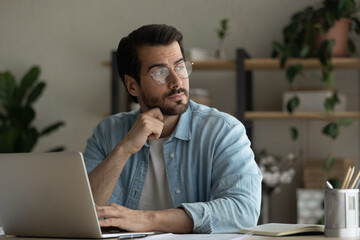 Thoughtful young business man in glasses sit at desk distracted from computer work looks into...