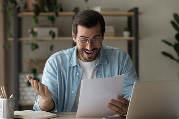 Fototapeta na wymiar Surprised guy read exciting news feels happy got job promotion letter looks shocked. Man in glasses sit at homeoffice desk hold document papers sheets excited by money refund, great sale offer concept