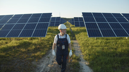 Adult engineer in uniform walking across solar battery panels on green sunny field. Futuristic...