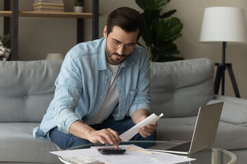 Serious millennial man in glasses calculates expenses working sit on sofa at home. Accountant job,...