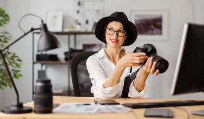 Professional female photographer reviewing photos on camera at office. Caucasian woman wearing trendy hat, white shirt and eyeglasses.