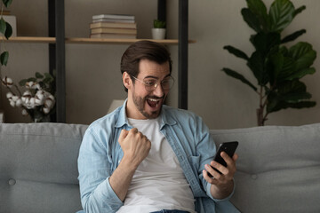 Excited overjoyed young man sit on sofa holding smartphone feeling euphoric with mobile online bet...