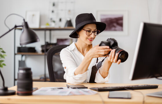 Positive caucasian woman in hat and eyeglasses sitting at desk with digital camera in hands. Professional photographer viewing taken photos at office.