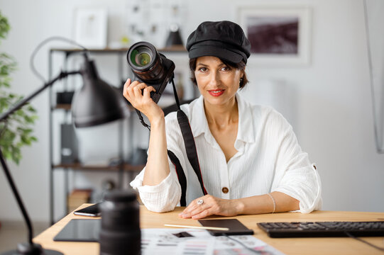 Portrait of beautiful woman in stylish wear sitting at modern office with digital camera in hands. Female photographer posing at workplace. Concept of creative hobby.