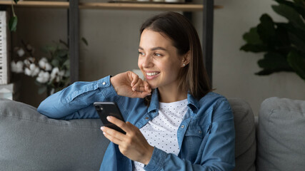 Attractive positive woman sit on sofa at home alone holding cell phone looking into distance. Modern wireless technology advanced user, generation addicted to tech, social media remote chat concept