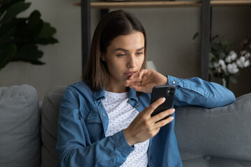 Serious thoughtful 25s woman sit on sofa at home holding smartphone device read sms feels upset,...
