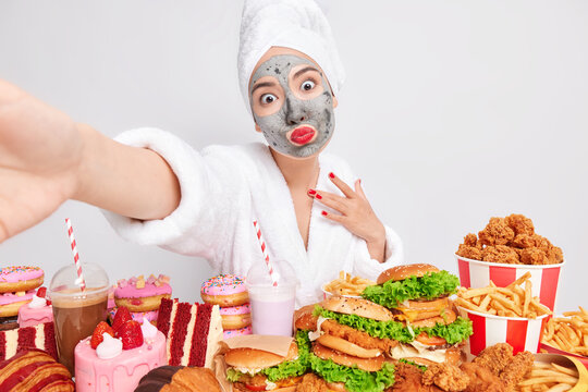 Romantive Asian woman applies clay mask on face to reduce fine lines keeps red lips rounded stretches arm for making selfie dressed in comfortable domestic clothes poses near delicious snacks