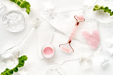 Pink quarts face roller, gua sha stone, pink cream in glass jar. Off white flat lay in green and pink with ice and exotic fern leaves. Beauty facial massage. Natural cosmetics laboratory.