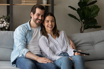 Obraz premium Laughing happy young couple hugging sit on cozy couch using remote controller switch channels watching internet television, funny tv show, comedy film, spend lazy weekend relaxing having fun at home