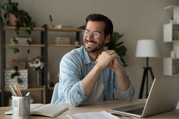 Smiling successful handsome man entrepreneur in glasses sit at desk looks into distance dreams about bright future, having business vision feels satisfied by career advance and personal growth concept