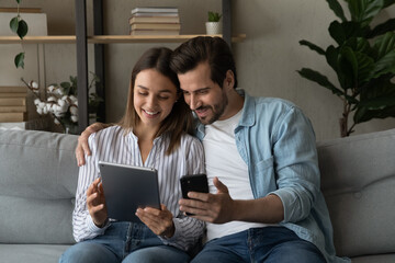 Young 30s couple sit on sofa use diverse gadgets holding smart phone and tablet device. Husband show explains to wife new mobile apps, comparing applications enjoy modern tech usage on weekend at home