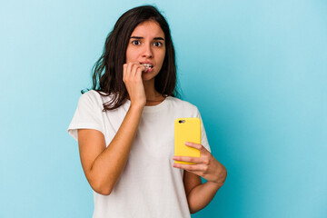 Young caucasian woman holding a mobile phone isolated on blue background biting fingernails, nervous and very anxious.