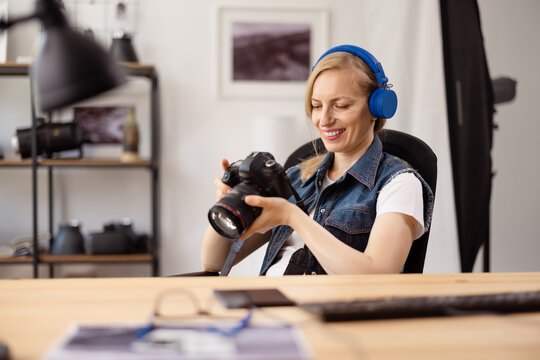 Smiling female photographer in wireless headphones sitting at table and holding digital camera in hands. Mature woman reviewing taken pictures at modern office.