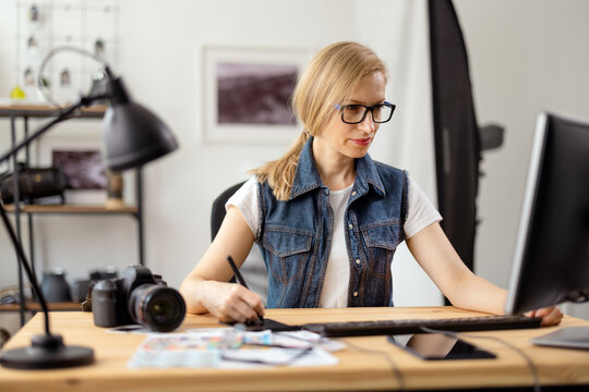 Focused photographer with blond hair editing taken pictures on modern computer. Caucasian woman in eyeglasses using various devices for work at office.