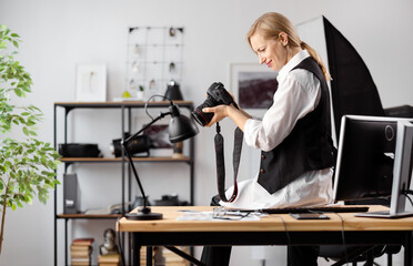 Side view of pleasant woman with blond hair sitting on table and adjusting photo camera. Female photographer in stylish wear enjoying working process at office.