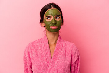 Young caucasian woman wearing a bathrobe and facial mask isolated on pink background confused, feels doubtful and unsure.