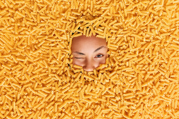 Mysterious young woman winks eyes raises eyebrows buried in scattering spiral pasta. Human face...