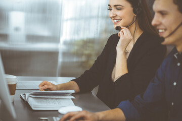 Young beautiful woman casual dressed in headsets is listening to a company's client, while she is sitting at the desk, working together with a male colleague in a modern office. Focus on woman. Call
