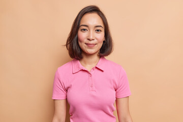 Portrait of young Asian female model with natural beauty dark hair rouge cheeks healthy skin looks happily at camera wears casual pink t shirt isolated over beige background. People concept.