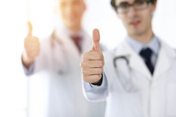 Two male doctors standing as a team and showing thumbs up as a symbol of the best service for patients in sunny clinic. Medicine concept