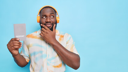 Thoughtful black bearded man holds chin looks away holds passport which contains basic identifying information listens music via wireless headphones isolated over blue background copy space area