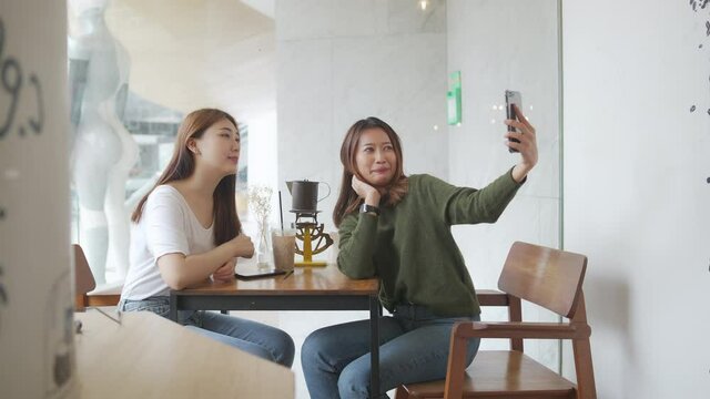 Two Asian woman friends sitting in cafe in the city and video calling on smartphone together. Female friendship enjoy and having fun outdoor lifestyle with wireless technology and modern gadget device