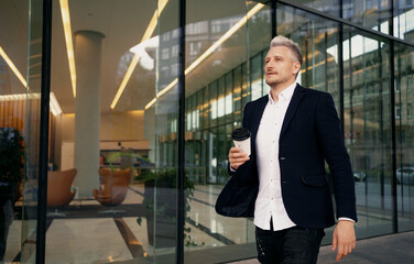 Fototapeta na wymiar He goes to work in a business center on the street. Good mood businessman. An adult male manager of European appearance is gray-haired, carries coffee in a paper cup in his hand.