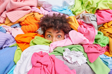 Surprised curly haired ethnic young woman stuck in pile of laundry does housework going to fold items. Human head in huge heap of unfolded clothes. Decluttering donation and housekeeping concept. - obrazy, fototapety, plakaty