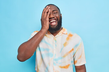 Upbeat cheerful bearded Afro American man makes face palm laughs happily expresses happy authentic emotions dressed in washed out t shirt isolated over blue background laughs at something funny
