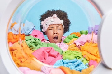Tired sleepy woman yawns after doing laundry buried in multicolored dirty clothes loaded in washer for washing. Busy housekeeper washes clothing at home. People housework and tiredness concept