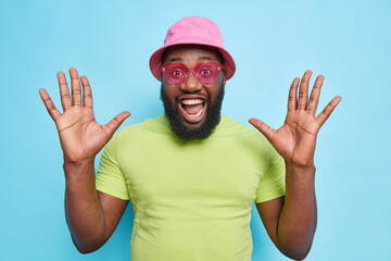 Naklejka premium Happy emotional dark skinned bearded guy raises palms feels very glad exclaims loudly reacts on amazing news wears stylish pink sunglasses casual t shirt and panama isolated over blue background