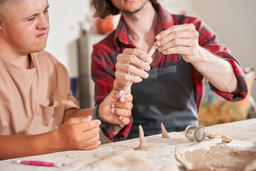 Man with special needs working with clay in ceramic studio under supervision of craftsman