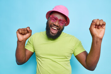Fototapeta premium Handsome positive dark skinned bearded man has fun on summer party dances carefree keeps arms raised up wears pink panama heart shaped sungasses and green t shirt isolated on blue studio wall