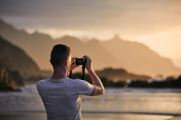 Rear view of man during photographing landscape with cliff. Photographer on beach at moody sunset. Tenerife, Canary Islands, Spain.