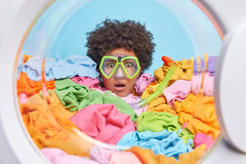 Impressed curly haired Afro American woman stares wondered through snorkeling glasses buried in multicolored clothes does laundry regularly. View from washing machine drum. Housekeeping routines