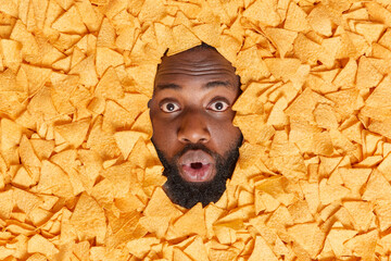 Impressed dark skinned bearded man shows face through heap of mexican chips keeps mouth opened from great wonder has thick beard cannot believe his eyes eats unhealthy snack. Harmful nutrition