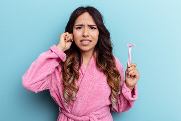 Young mexican woman wearing a bathrobe holding a knife isolated on blue background covering ears with hands.